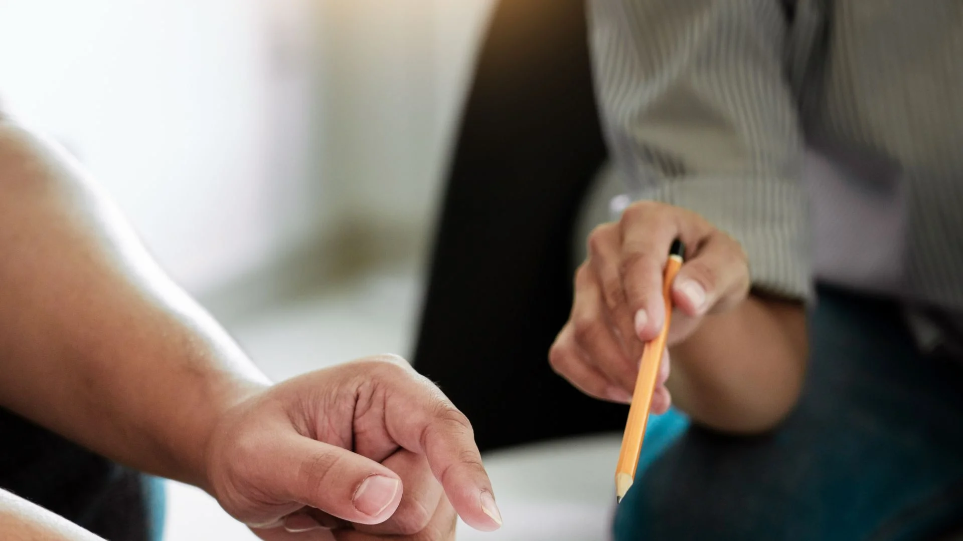 dłonie dwojga ludzi nad dokumentem, jedna dłoń wskazuje coś na dokumencie palcem, druga dłoń wskazuje coś trzymanym w ręku ołówkiem